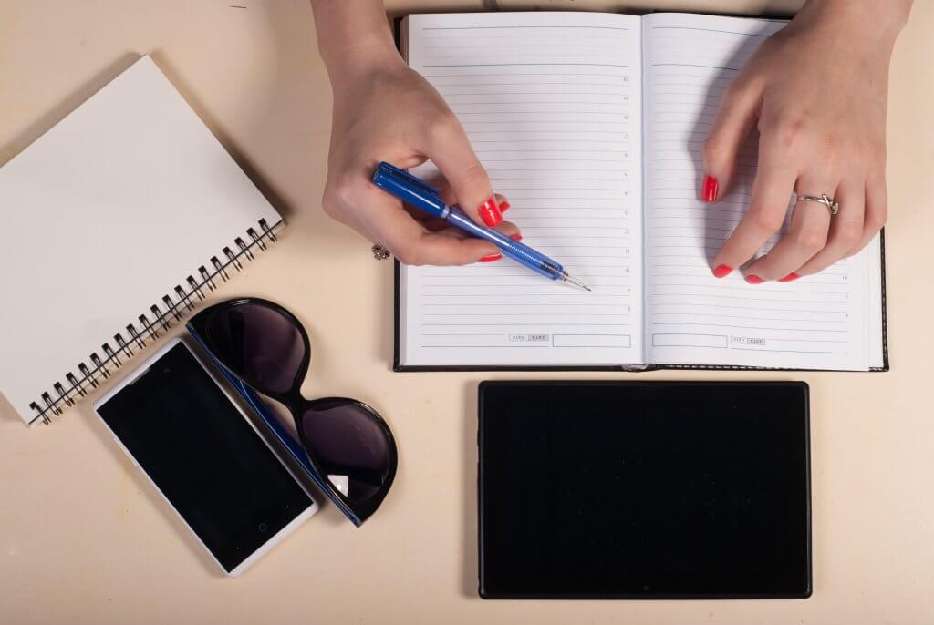 Start-Up Efficiency: 6 of the best free apps for a productive day. Photo - Woman writing in diary with a tablet, sunglasses and another notebook